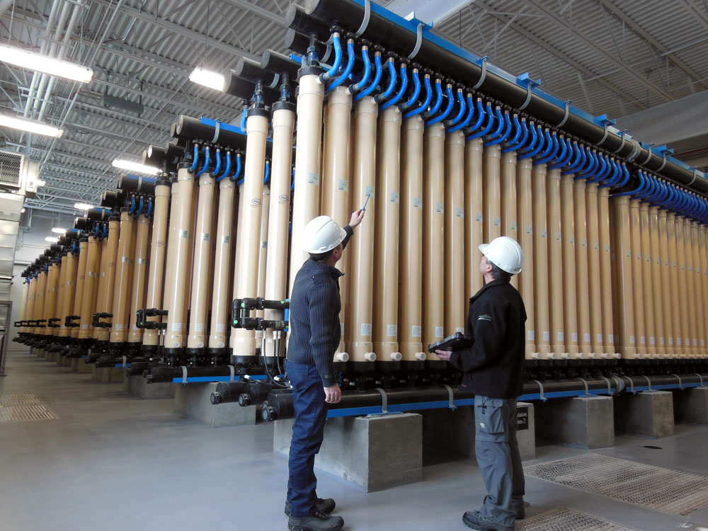 Two men in hard hats standing next to a large number of pipes.