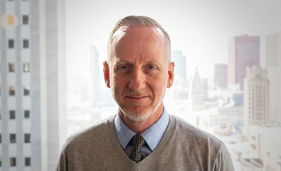 A man in a gray sweater standing in front of a city.