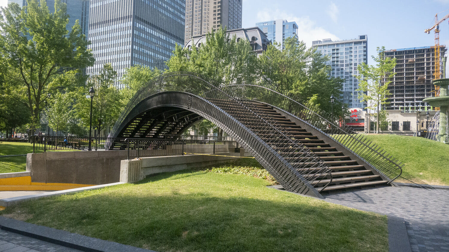 A bridge in the middle of a park with tall buildings in the background.
