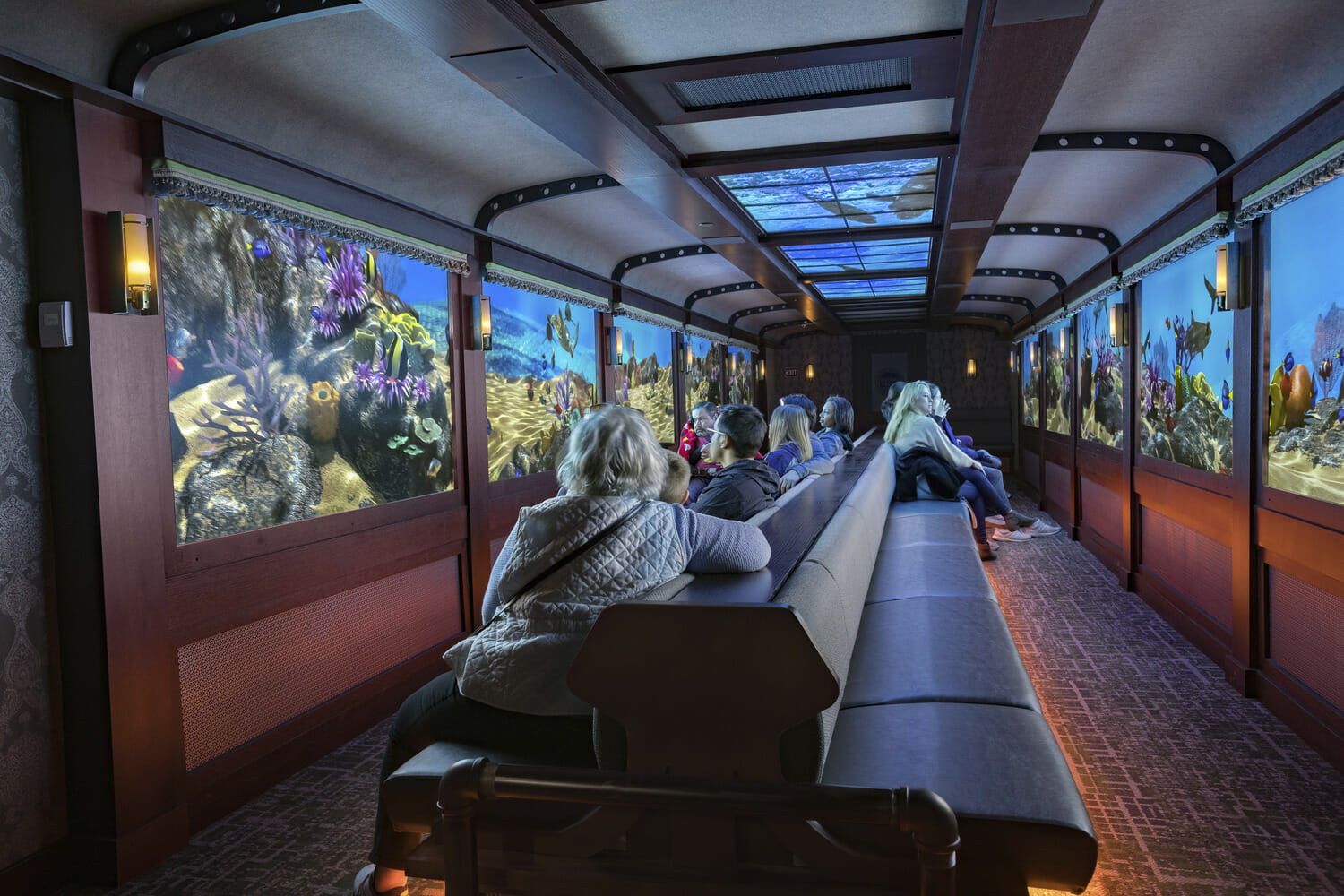 A group of people sitting on a train looking at a large screen.