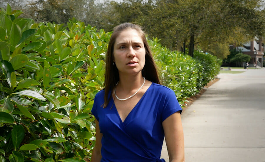 A woman in a blue dress standing in front of bushes.