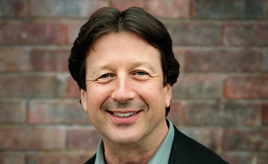 A man smiling in front of a brick wall.