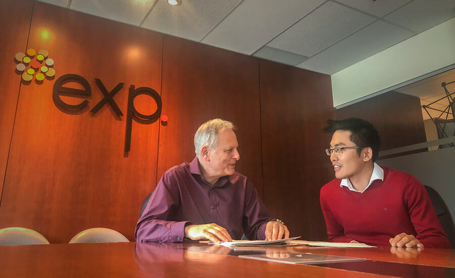 Two men sitting at a conference table with an exp sign in the background.