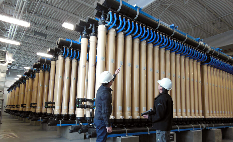 Two men in hard hats standing in front of a large number of pipes.