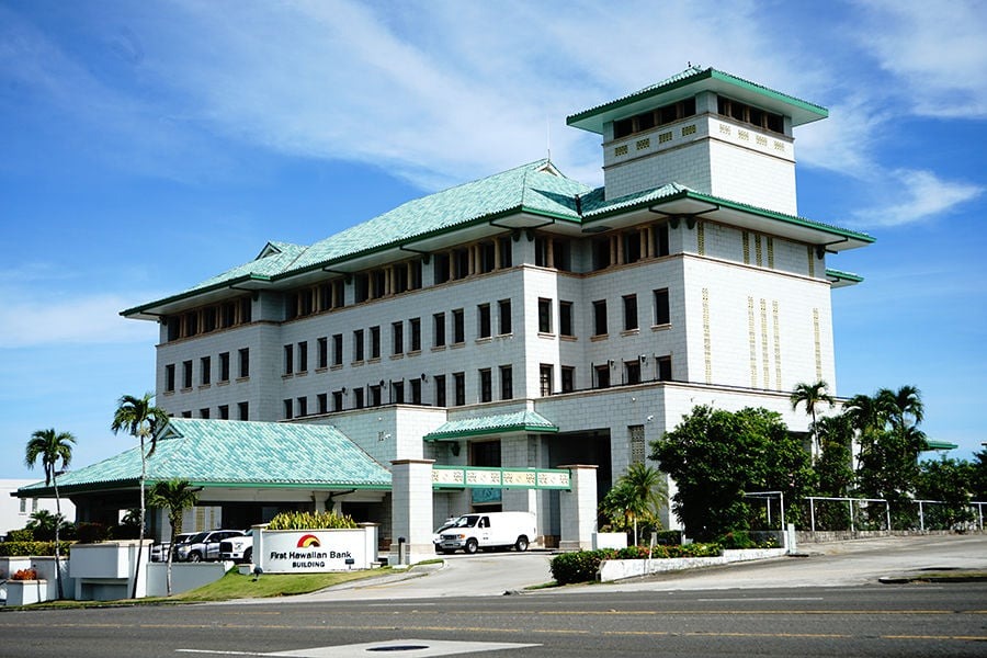 A large, multi-story building with a green-tiled roof, surrounded by palm trees and parked vehicles, identified as a bank by the sign reading “First Hawaiian Bank.”.