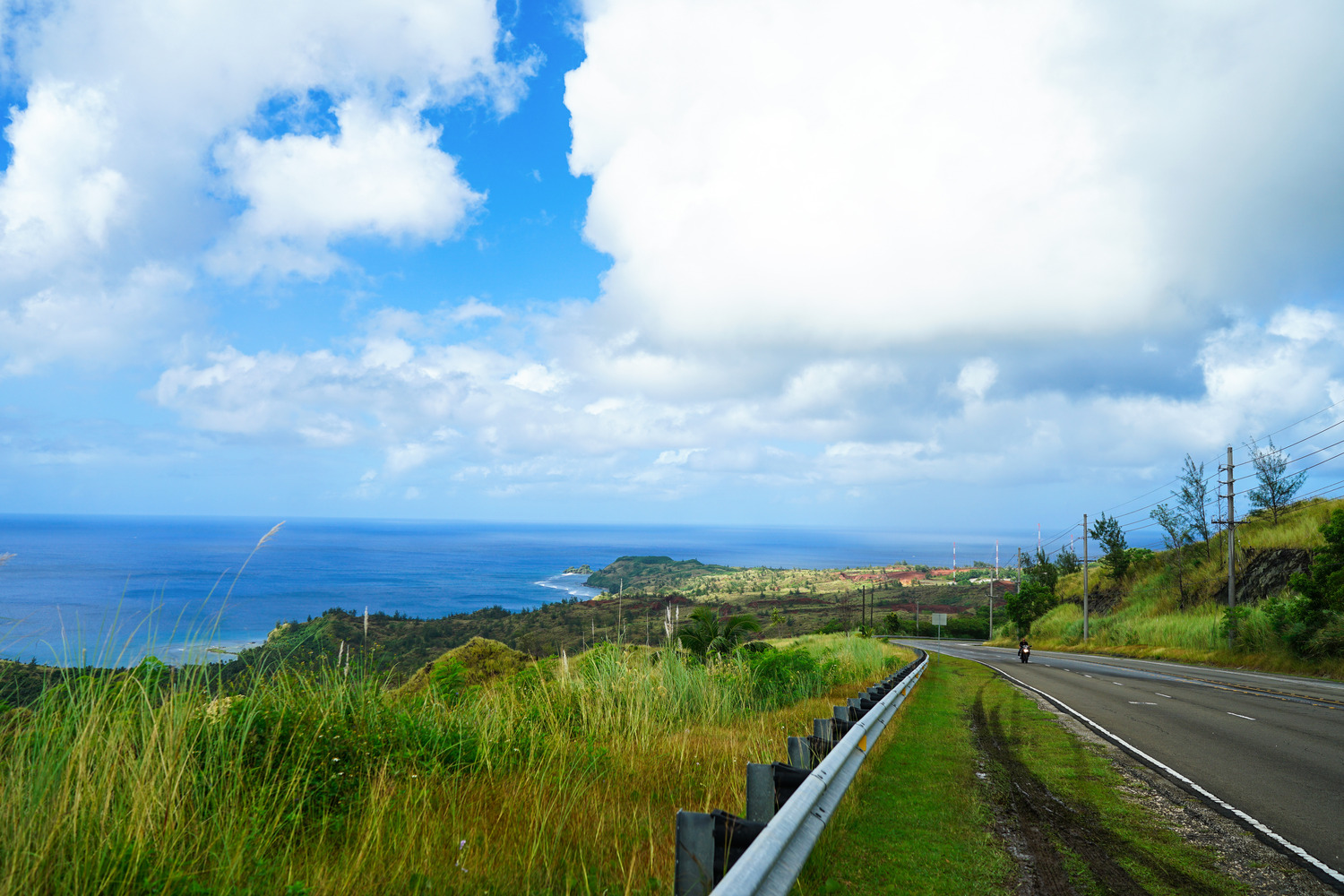 Une route pavée serpente le long d'une colline herbeuse surplombant l'océan sous un ciel bleu partiellement nuageux.