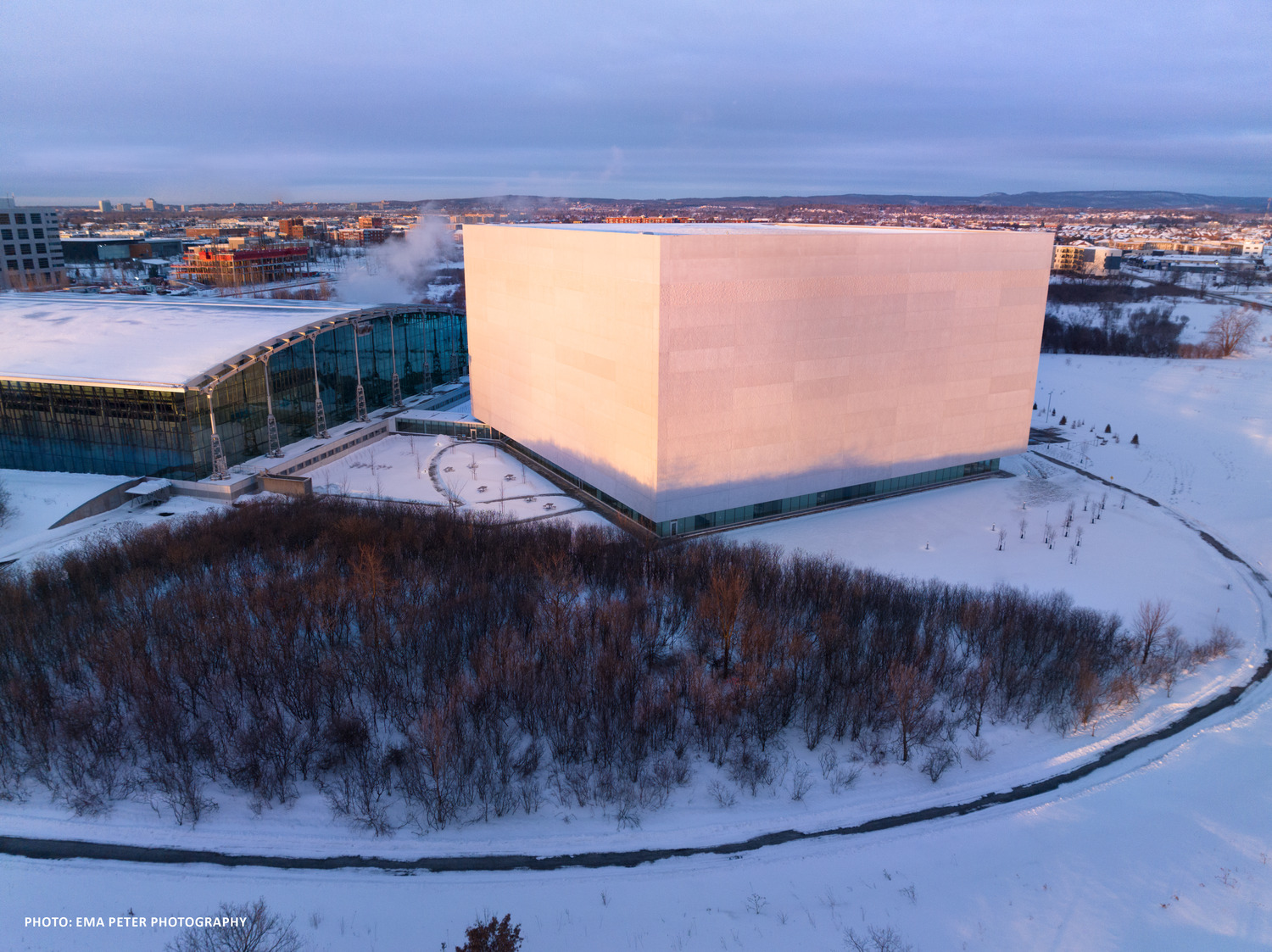 Vue aérienne d'un grand bâtiment cubique entouré d'un sol et d'arbres enneigés, avec un horizon de la ville en arrière-plan au crépuscule.