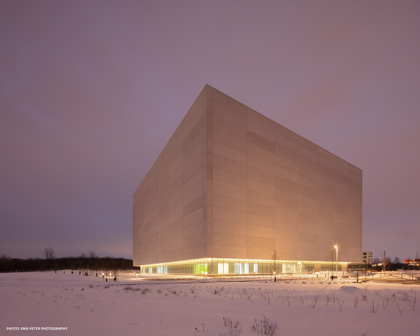 A modern, cube-shaped building illuminated at dusk, surrounded by a snowy landscape under a cloudy sky.