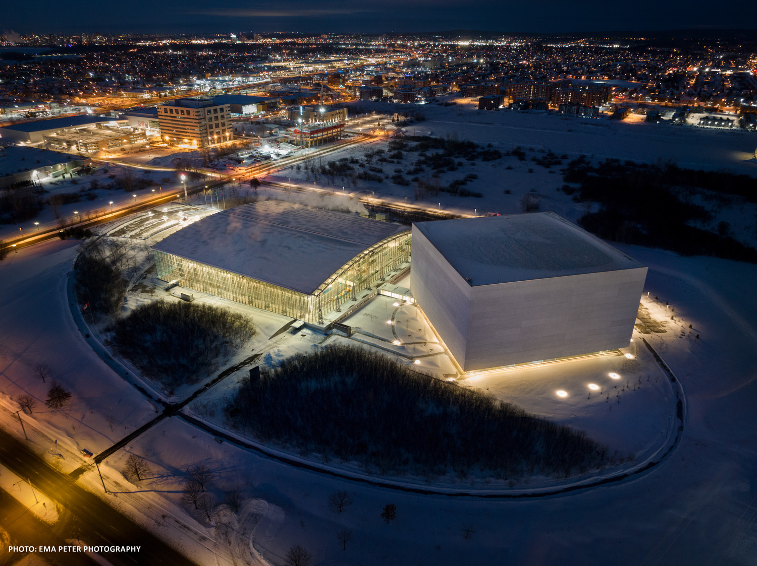 Vue aérienne d'un bâtiment moderne avec des murs de verre éclairés et une grande structure de cube blanc à côté, entouré d'un paysage enneigé la nuit.