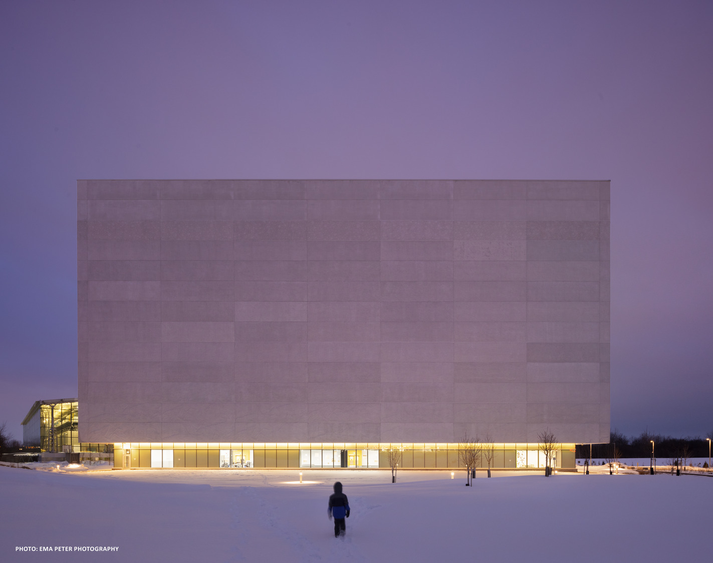 An illuminated modern building with a large, flat facade on a snowy landscape during twilight, with a person approaching on a footpath.