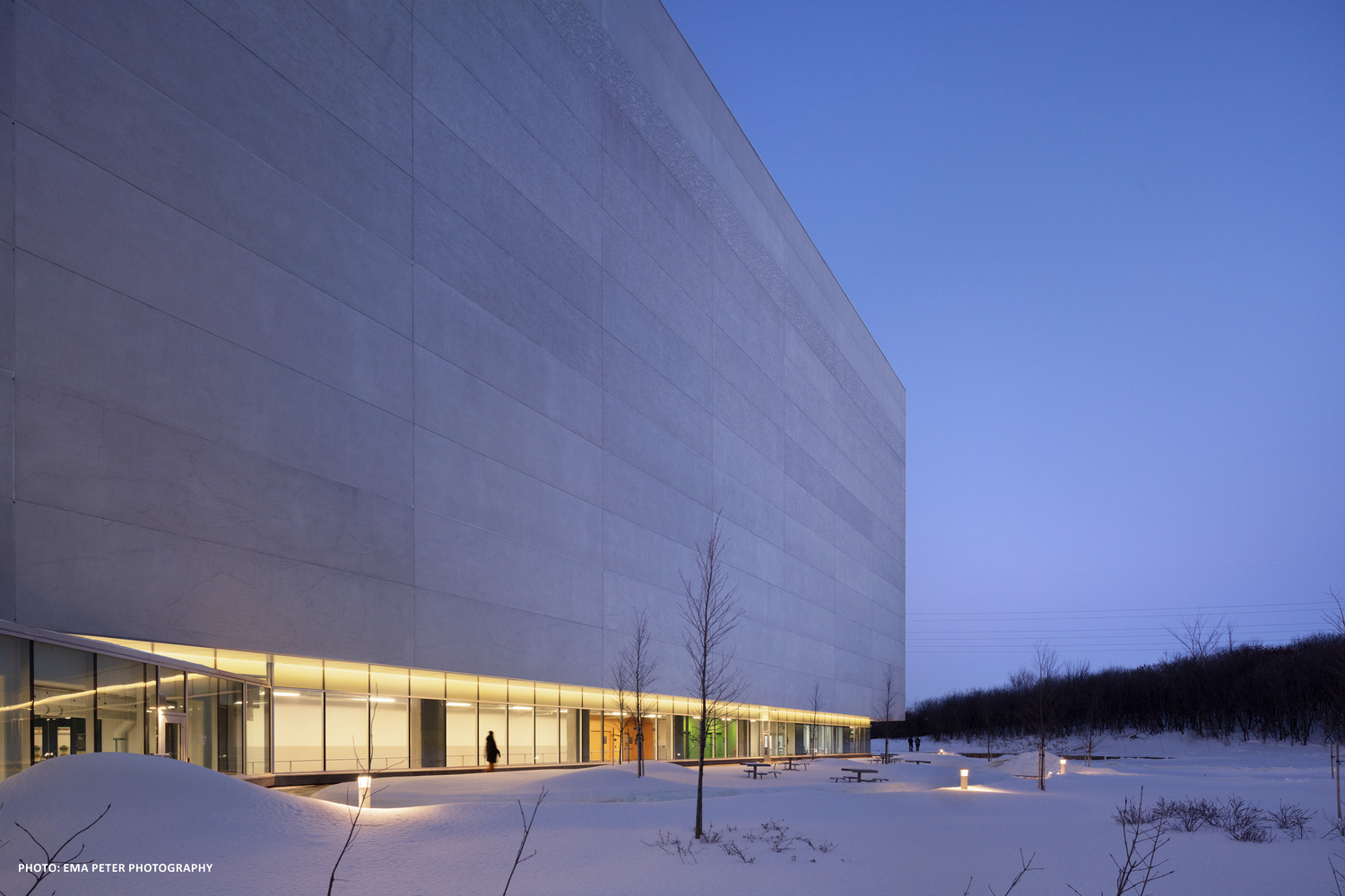 Extérieur d'un grand bâtiment moderne avec fenêtres éclairées au crépuscule, entouré d'un paysage enneigé.