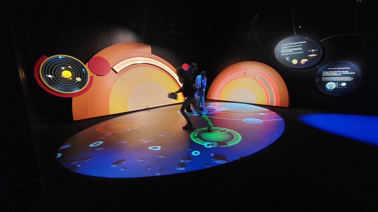 Deux personnes traversent une salle obscure dont les murs et le sol sont couverts d'écrans interactifs circulaires et colorés et de projections liées à l'espace.