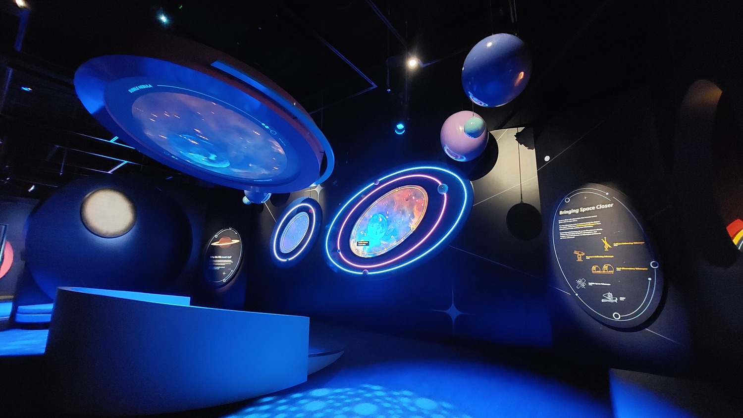 Une salle d'exposition sombre présente des écrans circulaires éclairés et des modèles planétaires suspendus, qui fournissent des informations sur l'espace et les corps célestes.