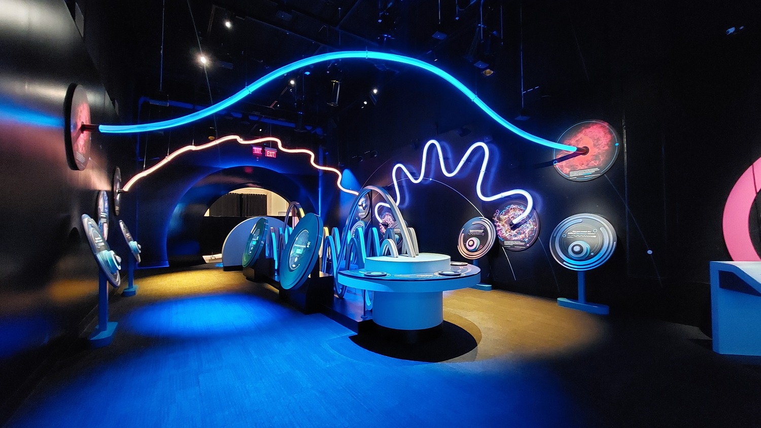 Une exposition dans un musée scientifique moderne avec des lumières bleues brillantes, des écrans interactifs et des écrans circulaires sur des murs sombres, avec une personne assise à une table centrale.
