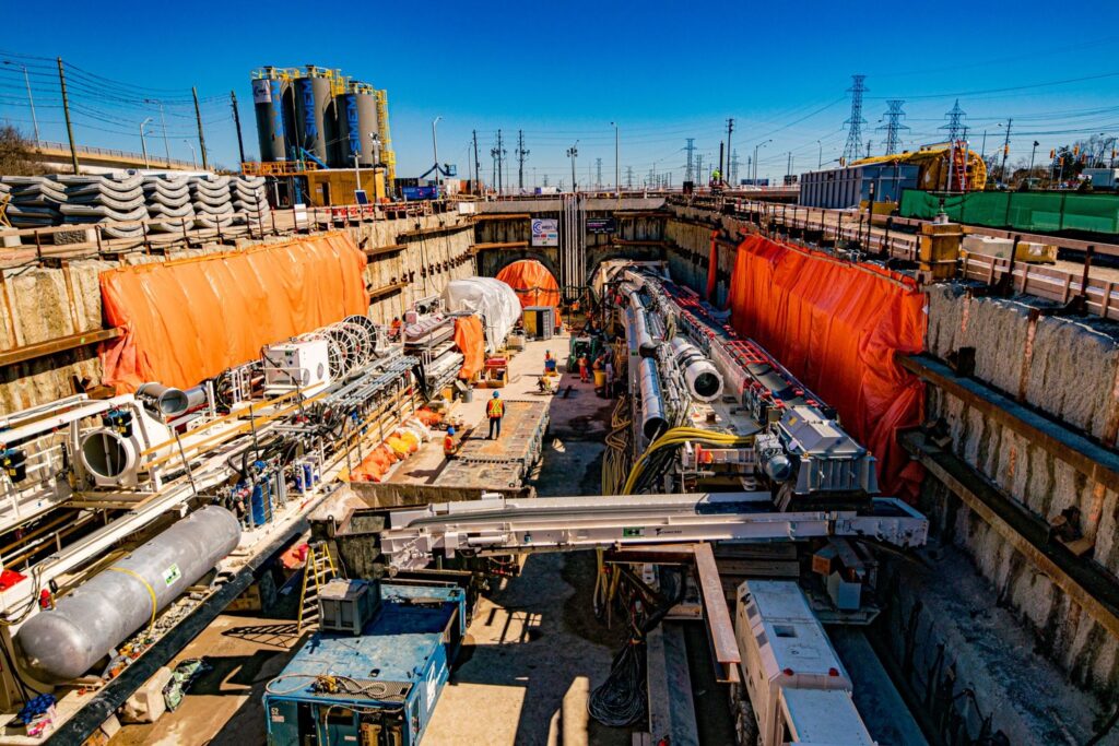 Construction site featuring workers and machinery engaged in a large-scale tunneling project. Equipment and materials are organized along both sides of the trench, with tall structures in the background.