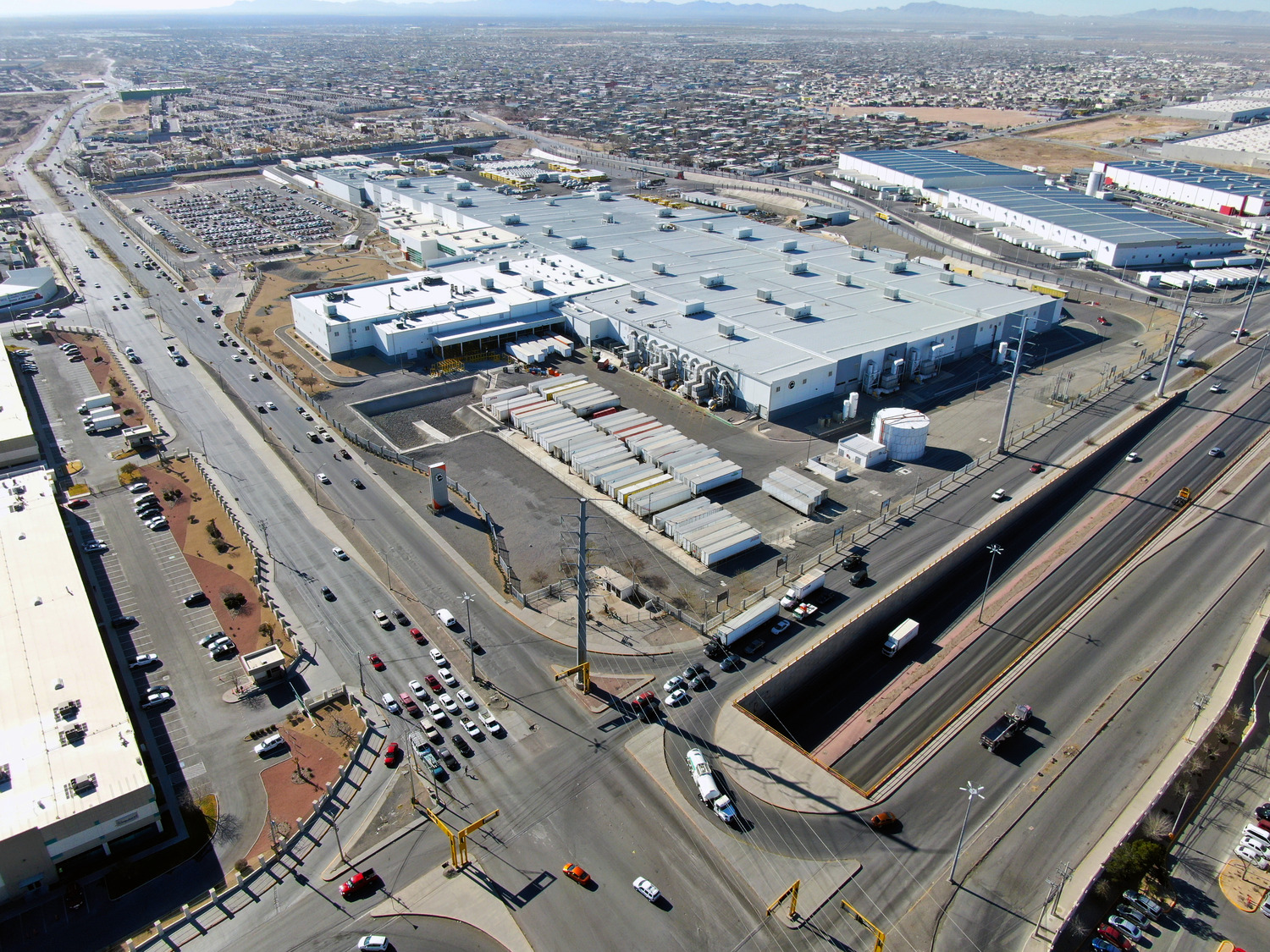 Vue aérienne d'un grand complexe industriel à côté d'une autoroute à plusieurs voies et à circulation modérée, entouré d'une zone suburbaine avec de nombreux bâtiments.