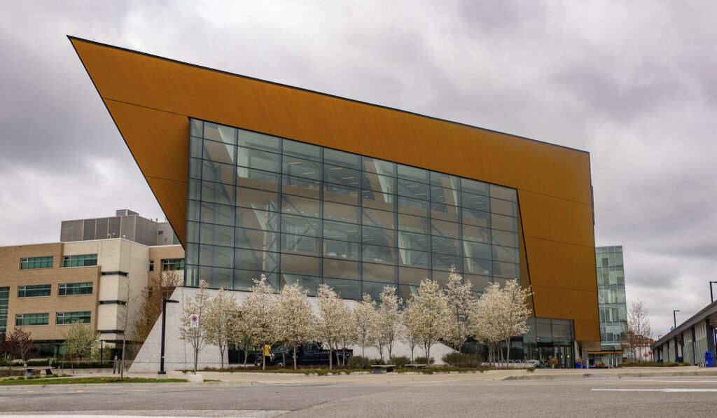 Modern angular building with large glass facade and blooming trees in the foreground under overcast skies.