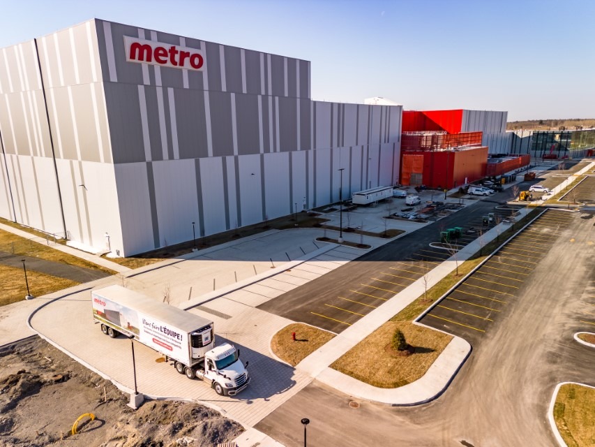 Vue aérienne d'un centre de distribution de métro avec un camion de livraison stationné à l'extérieur, entouré d'un parking pavé et d'un chantier de construction adjacent.