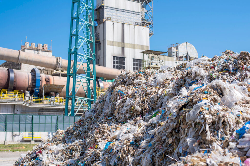 A large pile of mixed waste in the foreground with an industrial recycling facility in the background on a clear day.