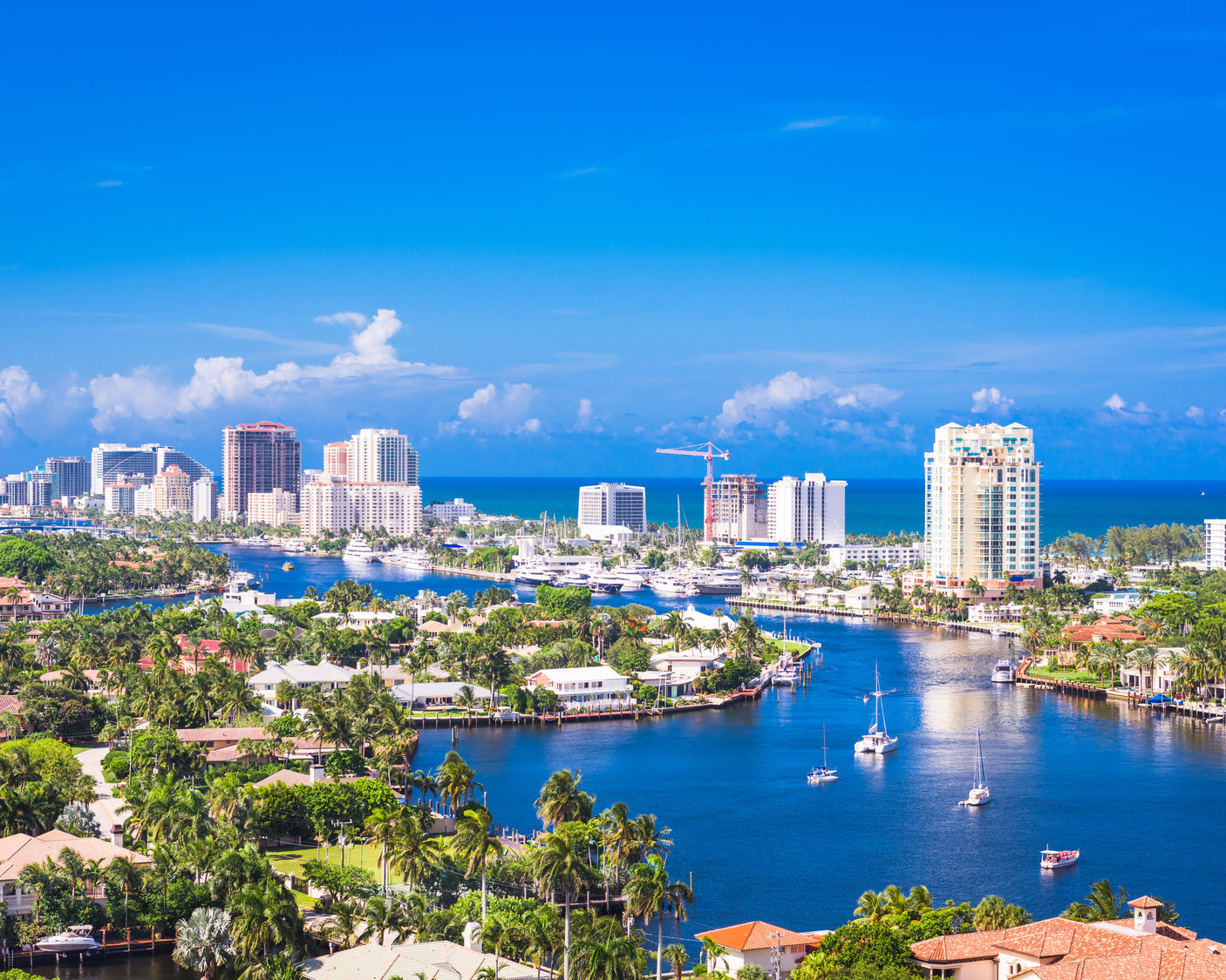 Aerial view of a coastal city with high-rise buildings, a winding river, boats, and lush greenery on a sunny day with clear skies.