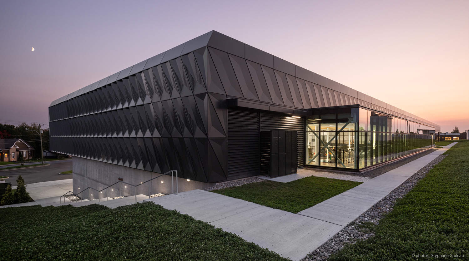 Immeuble de bureaux moderne avec une façade en verre réfléchissante et des panneaux métalliques géométriques au crépuscule, avec une allée paysagée et un croissant de lune dans le ciel.