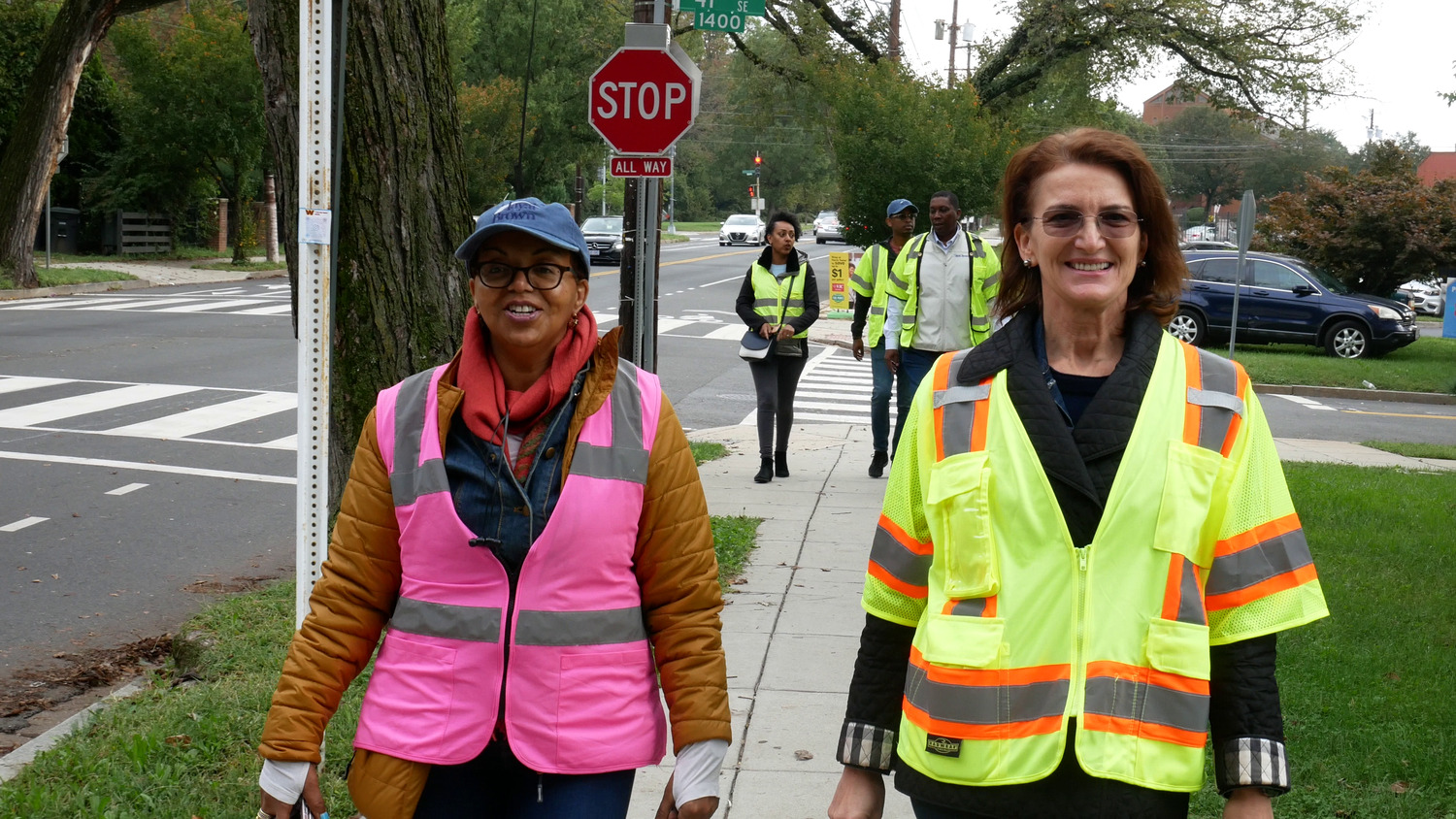 Two women wearing safety vests smile while walking on a sidewalk, with other pedestrians and a stop sign in the background.