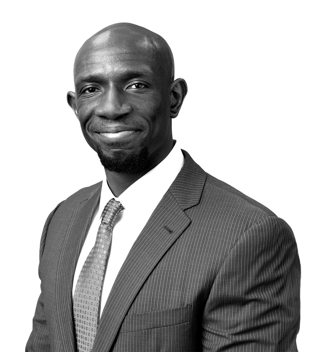 Headshot of a smiling bald man in a pinstriped suit and patterned tie, portrait styled in black and white.