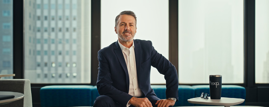 A professional man in a suit sits on a table in a modern office with large windows and a cityscape view.