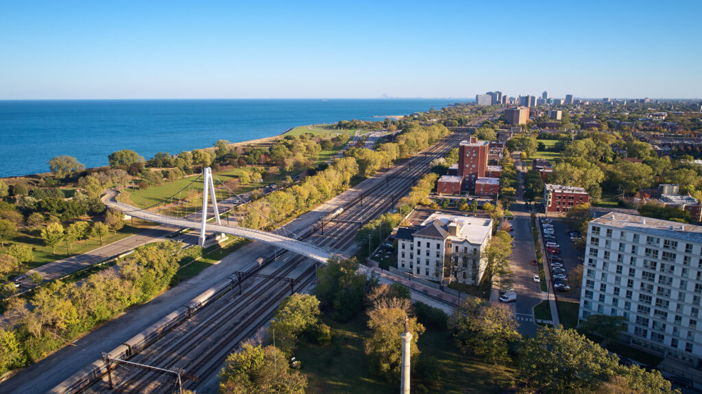 Aerial view of a green urban area beside a body of water, featuring a footbridge over railroad tracks, residential buildings, and a distant city skyline.