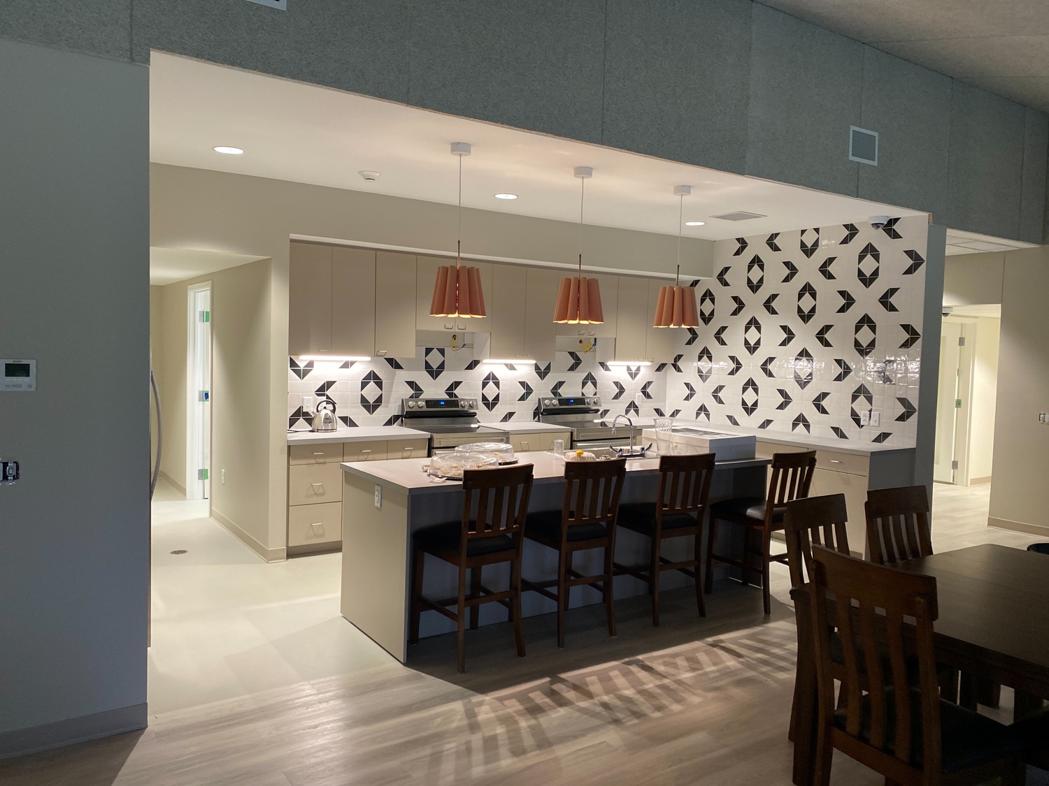 Open modern kitchen with an island featuring four bar stools, pendant lights, and a geometric backsplash. Dining table with chairs visible in the foreground. Walls and floor are light-colored.