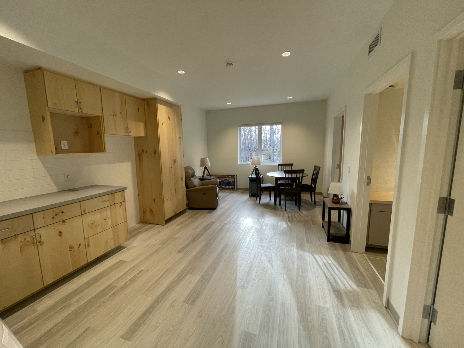 A brightly lit, modern apartment with light wood flooring, wooden cabinetry on the left, a seating area, and a dining table near a window in the background. A doorway on the right leads to another room.