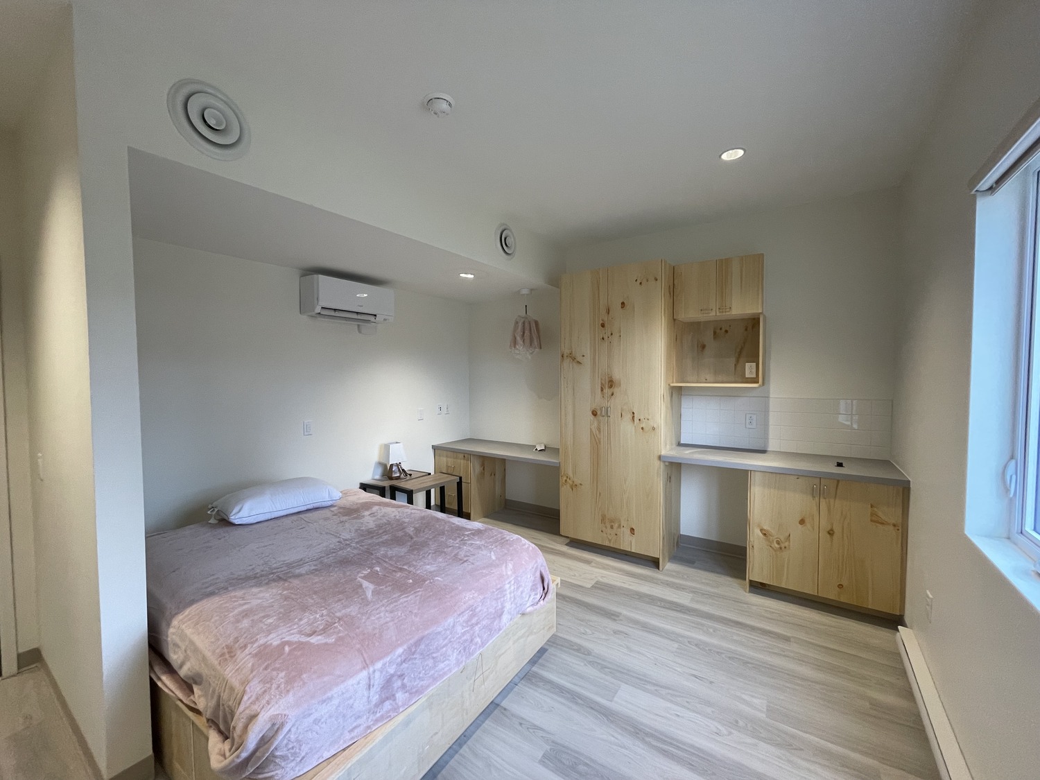 Une chambre minimaliste avec un lit soigneusement fait, un bureau en bois et des armoires. La chambre dispose de parquet en bois clair, de murs blancs et d'une petite kitchenette avec comptoir.