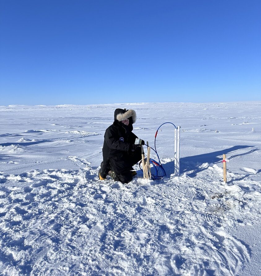 Person in a winter coat kneeling on snow-covered ground in a vast, flat landscape with clear blue sky, working with a small, makeshift setup involving tubes and wooden stakes.