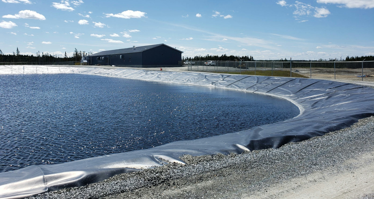 Un grand réservoir d'eau bordé d'ondulations sous un ciel clair, adjacent à une zone clôturée avec un long bâtiment industriel gris en arrière-plan.