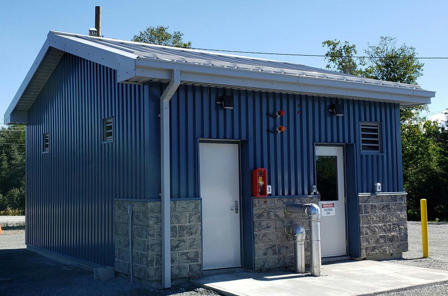 Petit bâtiment utilitaire en métal bleu et béton avec un toit en pente, deux portes et un extincteur rouge fixé au mur extérieur. Des arbres sont visibles en arrière-plan.