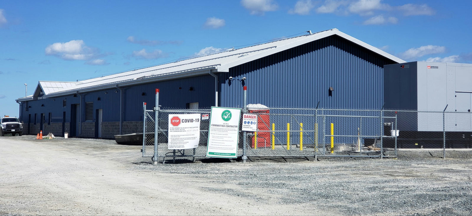 Un grand bâtiment industriel bleu avec un périmètre clôturé. Des panneaux sur la clôture comprennent des avertissements COVID-19 et des informations de sécurité. Un camion et du matériel de construction se trouvent à proximité.