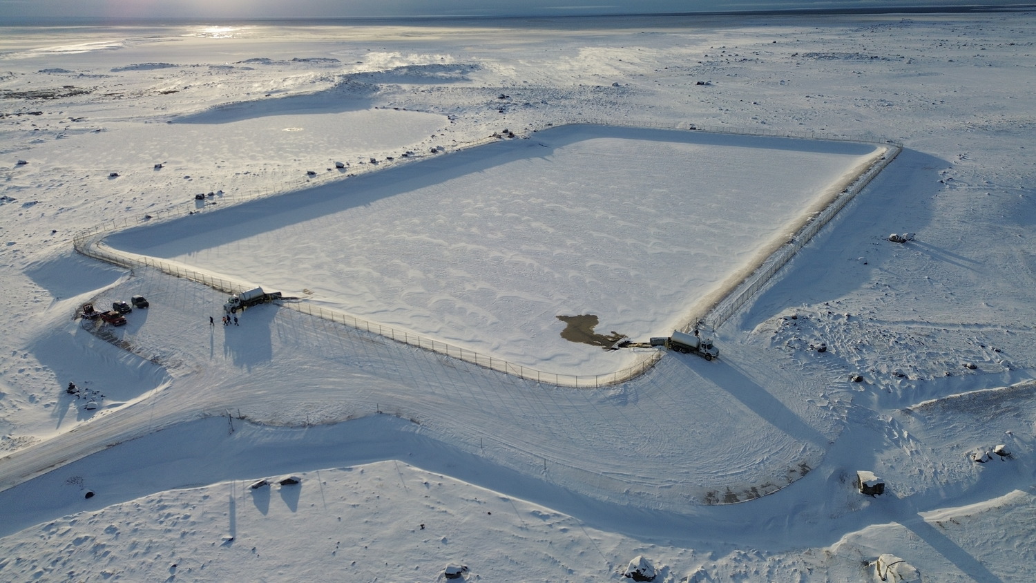 Aerial view of a large rectangular snow-covered area with surrounding fences in a snowy landscape. There are vehicles parked at one end.