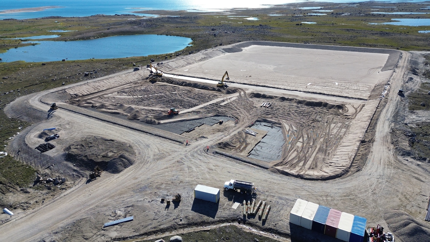 Aerial view of a construction site with machinery working on dirt areas near a body of water. Tents and equipment are visible in the foreground.