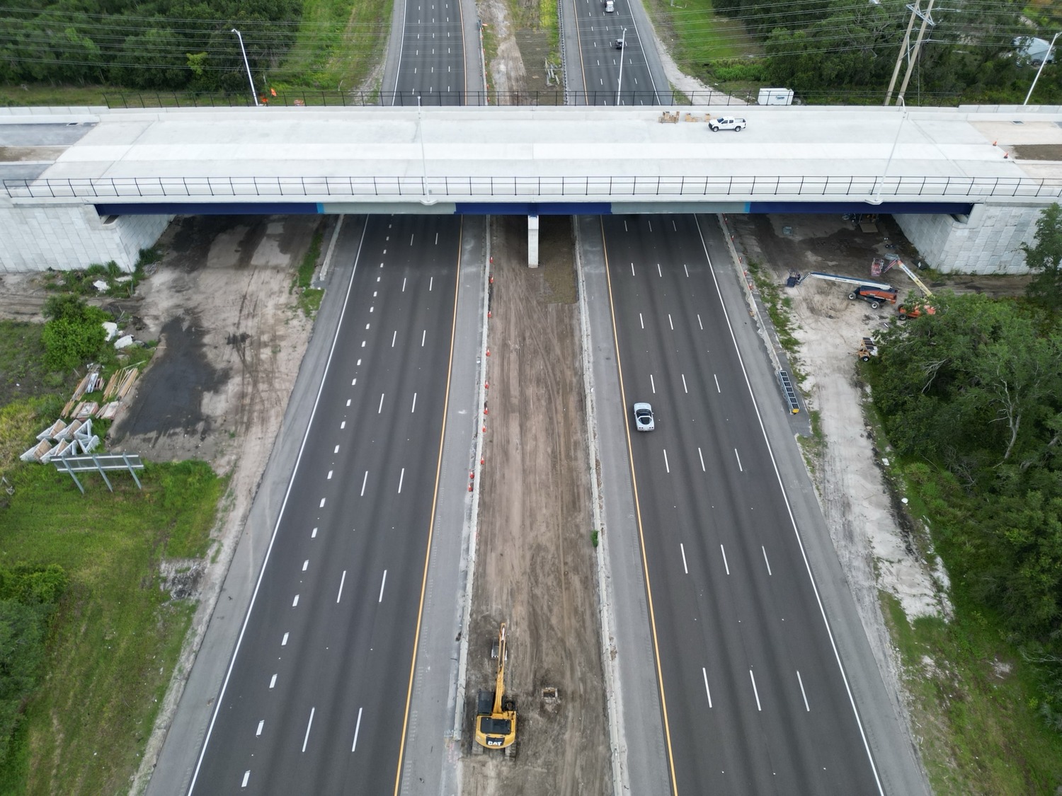 Vue aérienne d'une autoroute sous un viaduc, avec un véhicule de construction et de la terre sur la voie centrale, et une circulation légère sur les voies adjacentes.