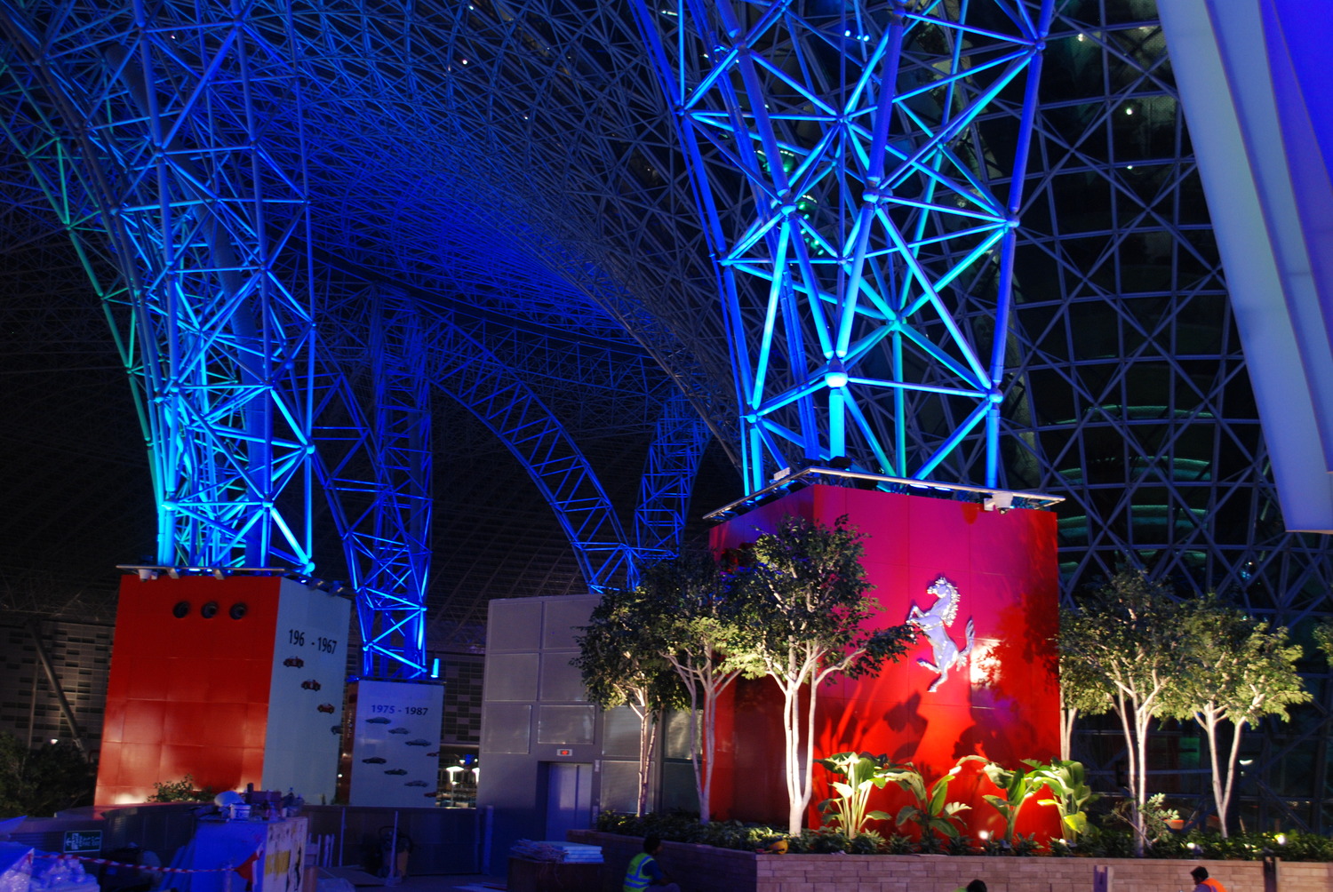 Grande structure intérieure avec poutres en acier éclairées en bleu et mur rouge avec logo représentant un cheval cabré. Des plantes luxuriantes entourent la base et une vue partielle d'un toit imposant est visible.