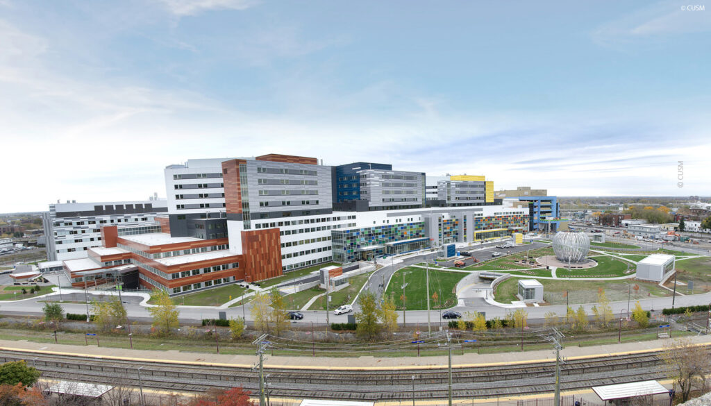 Panoramic view of a large, modern hospital complex with multiple connected buildings, green spaces, and surrounding roads and railway tracks.