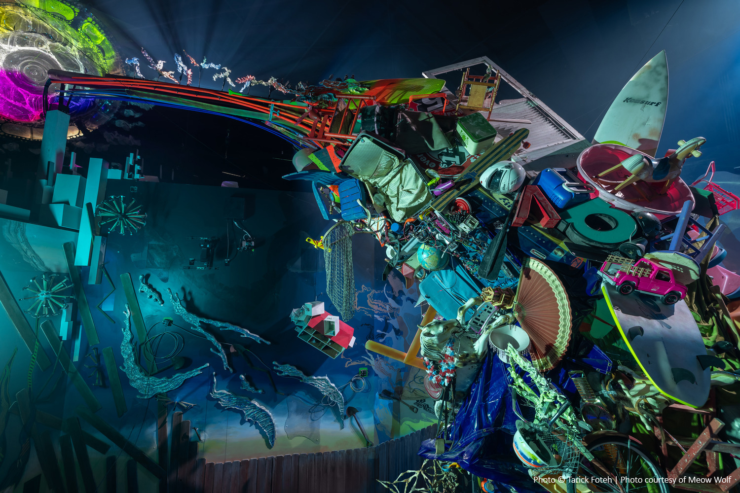 Une sculpture vibrante et chaotique composée d'objets et de matériaux variés, avec un éclairage coloré mettant en valeur ses détails et textures complexes sur un fond sombre.