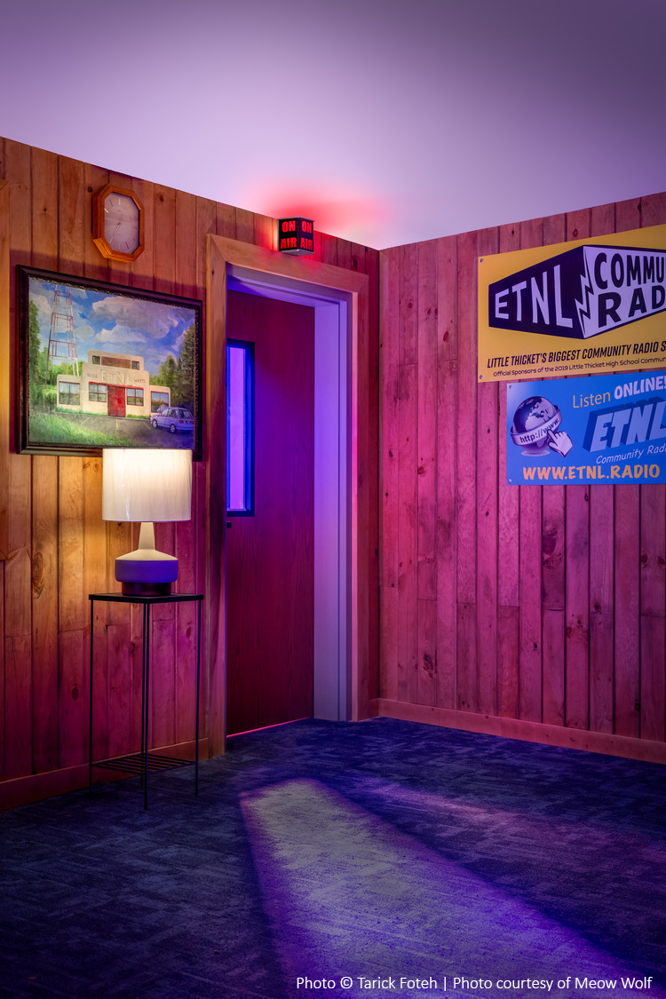 Une pièce aux murs lambrissés, avec un tableau, une lampe et des affiches pour la radio communautaire ETNL. Un panneau rouge « On Air » est illuminé au-dessus d'une porte partiellement ouverte.