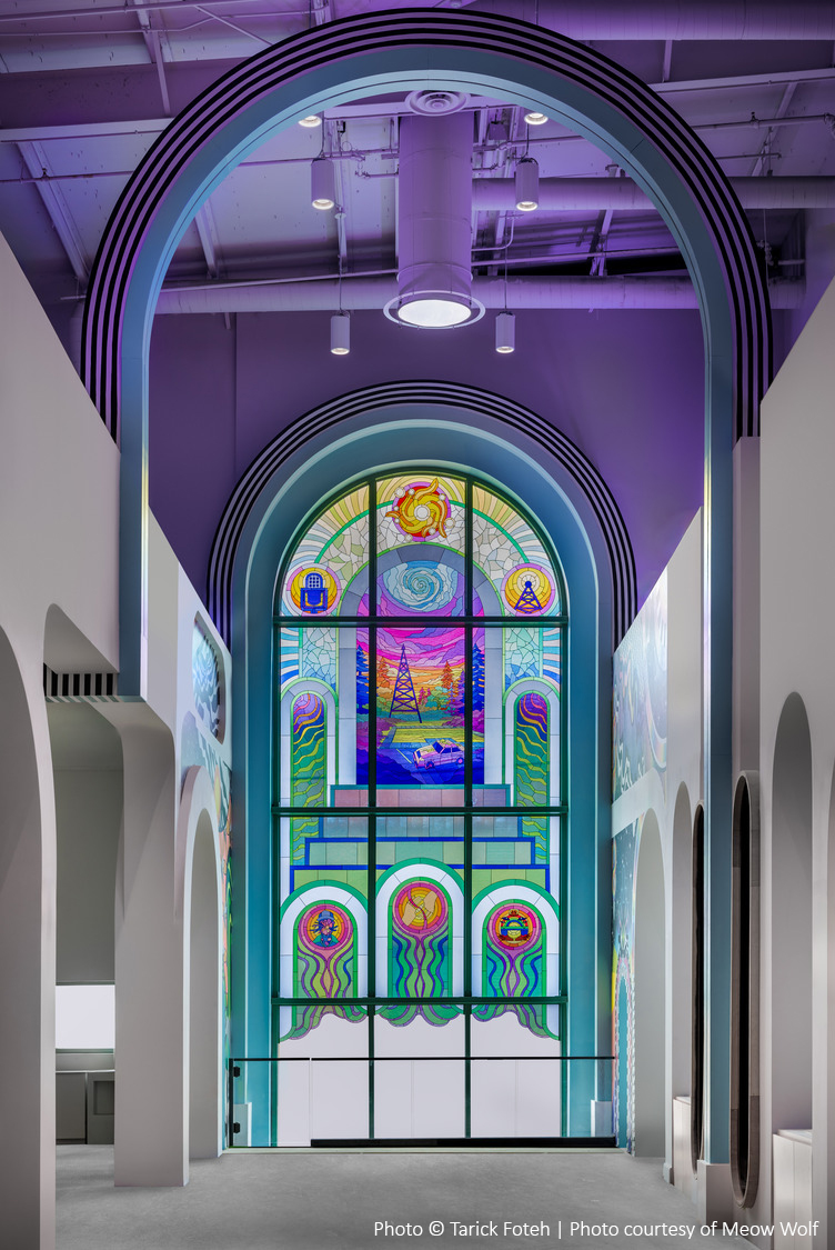 Un grand vitrail aux motifs abstraits colorés est encadré par une série d'arcades et est éclairé par des plafonniers.