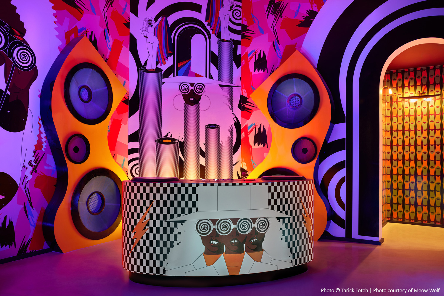Salle dynamique et futuriste avec de grandes enceintes, des œuvres d'art murales vives et une console cylindrique présentant des motifs abstraits et un motif en damier.