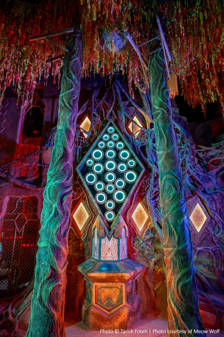 Une installation vibrante et surréaliste composée de lumières circulaires lumineuses disposées en forme de losange, entourées de piliers colorés et texturés, avec un plafond orné d'éléments suspendus.