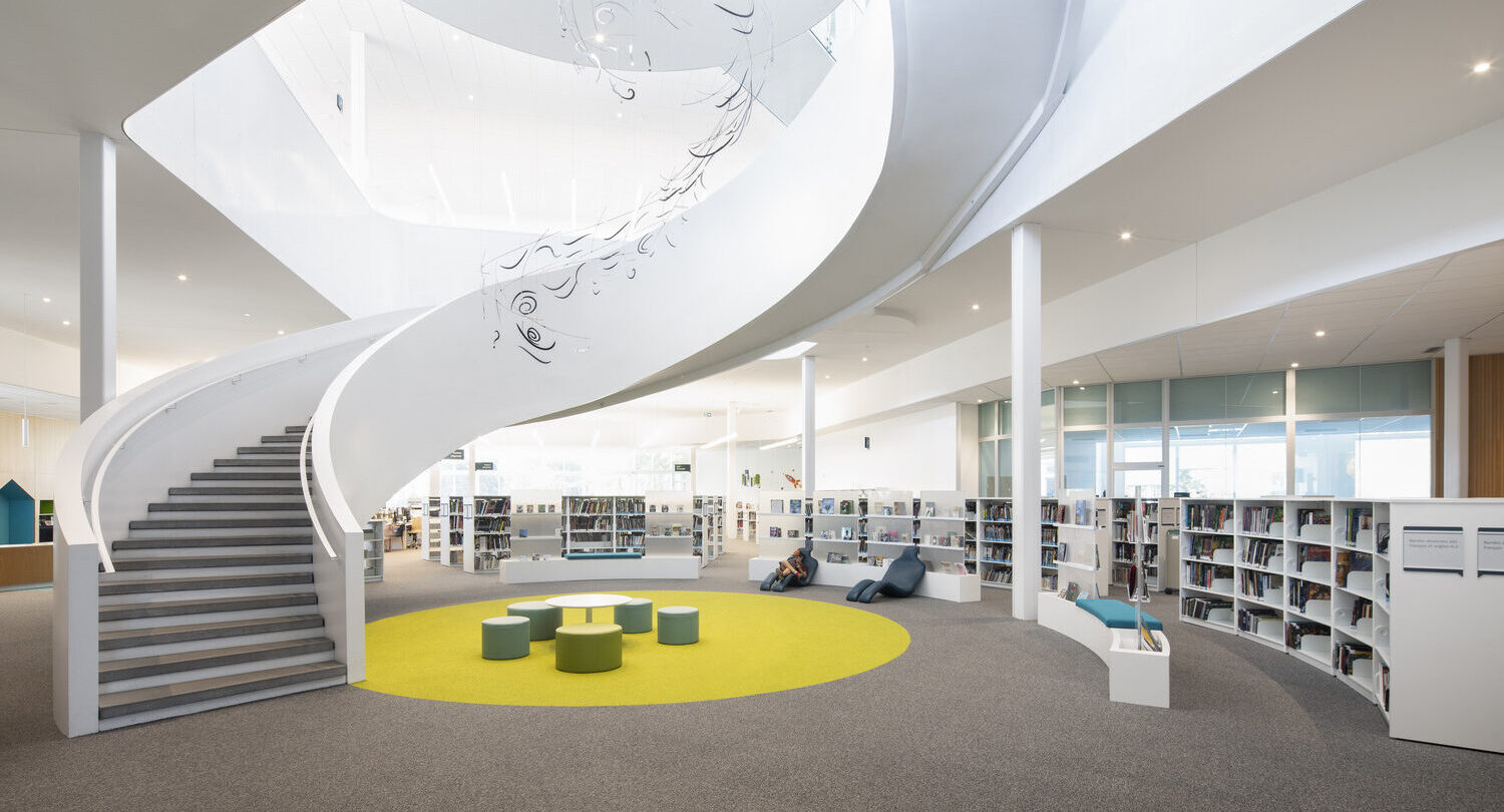 Intérieur de bibliothèque moderne avec escalier en colimaçon, coin salon circulaire sur tapis jaune et étagères. Éclairage lumineux et aménagement d'espace ouvert.
