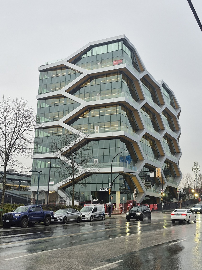 A modern, multi-story office building with a geometric, honeycomb-like facade showcases innovative building science and envelope design on a rainy day, as cars pass along the wet street in front.