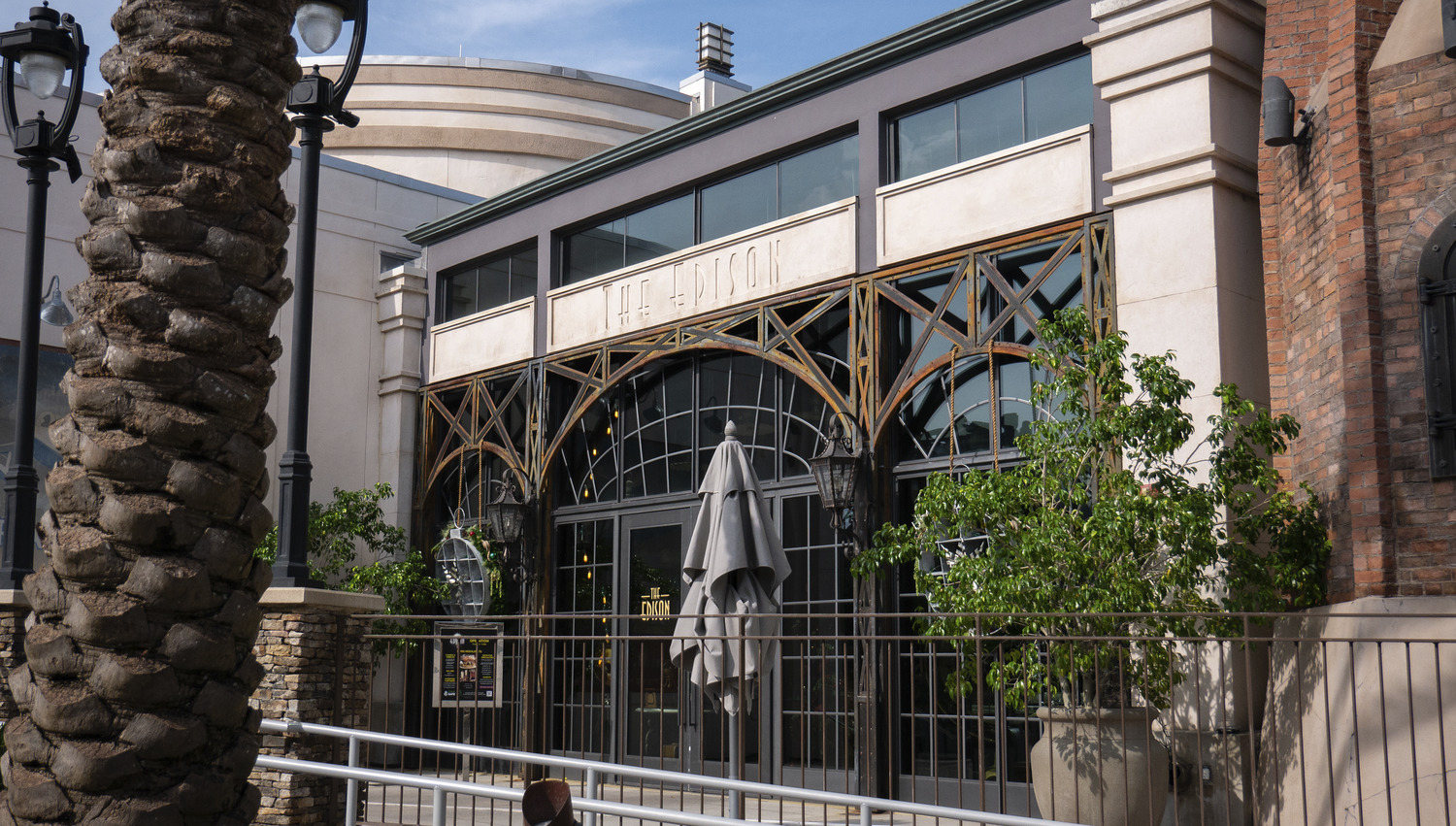 Exterior of a building with arched windows, ornate metalwork, and an umbrella closed on the patio. A palm tree and lamppost are visible in the foreground.