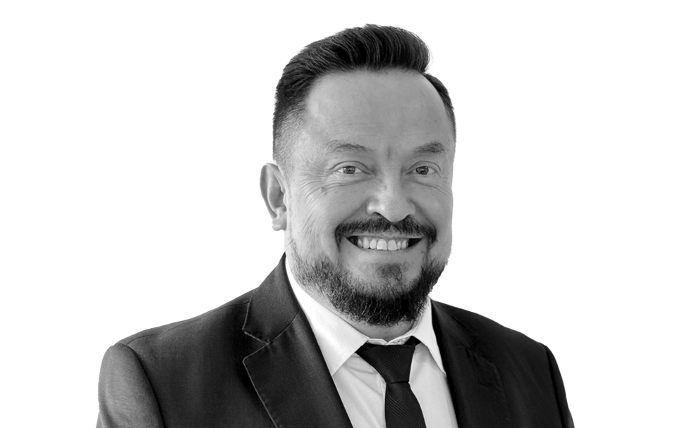 Smiling man in a suit and tie, with a beard and short hair, against a plain background.
